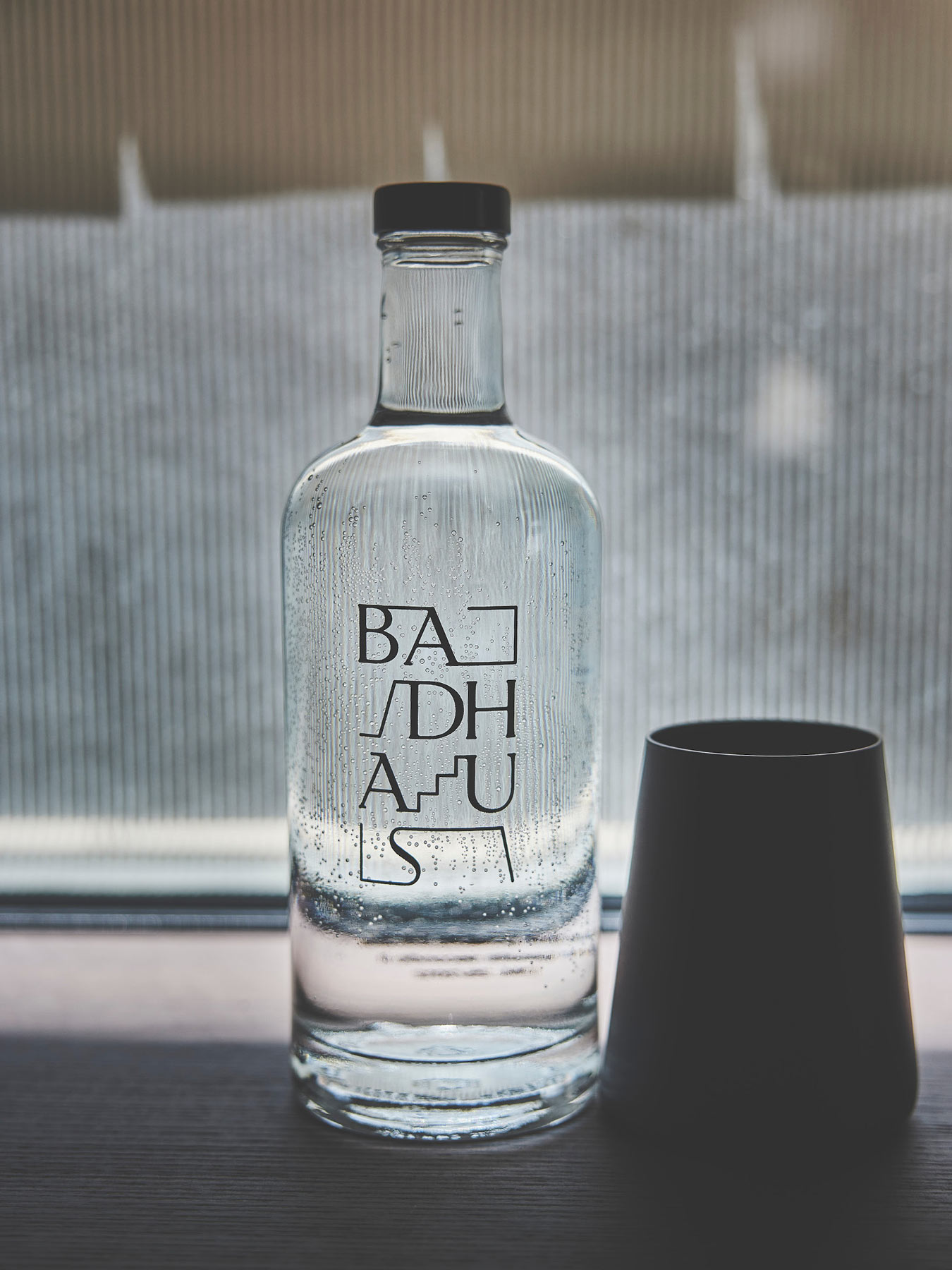 Wasserflasche mit Badhaus-Logo neben dunklem Becher auf Fensterbank