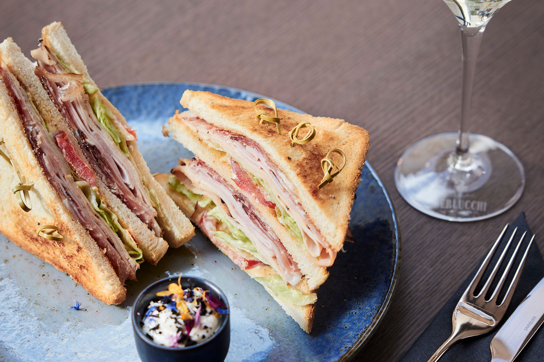 Club-Sandwich auf blauem Teller mit kleinem Dip und Weinglas