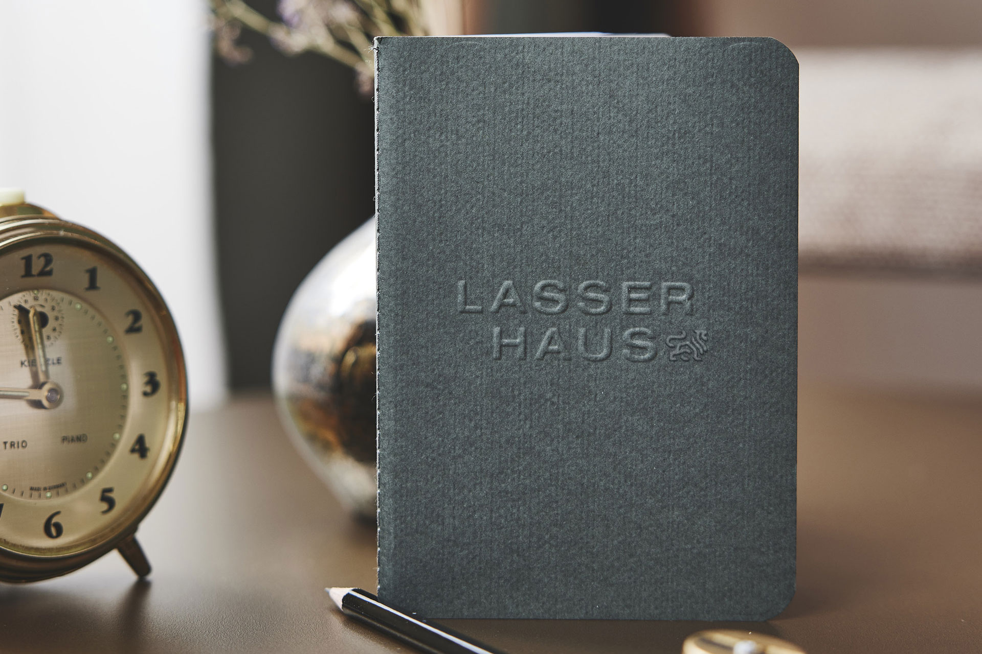 Notizbuch mit geprägtem 'LASSER HAUS' Titel, neben antikem Wecker und Kugelschreiber