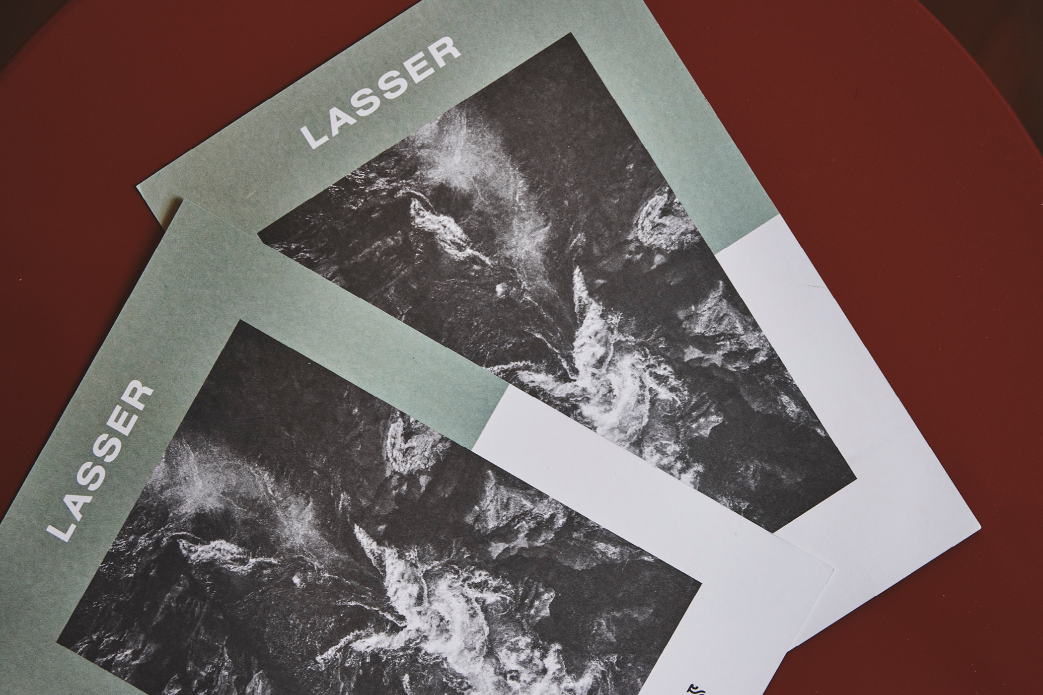 Zwei identische Flyer mit dem Schriftzug 'LASSER' und abstraktem Schwarz-Weiß-Motiv auf rotem Tisch.