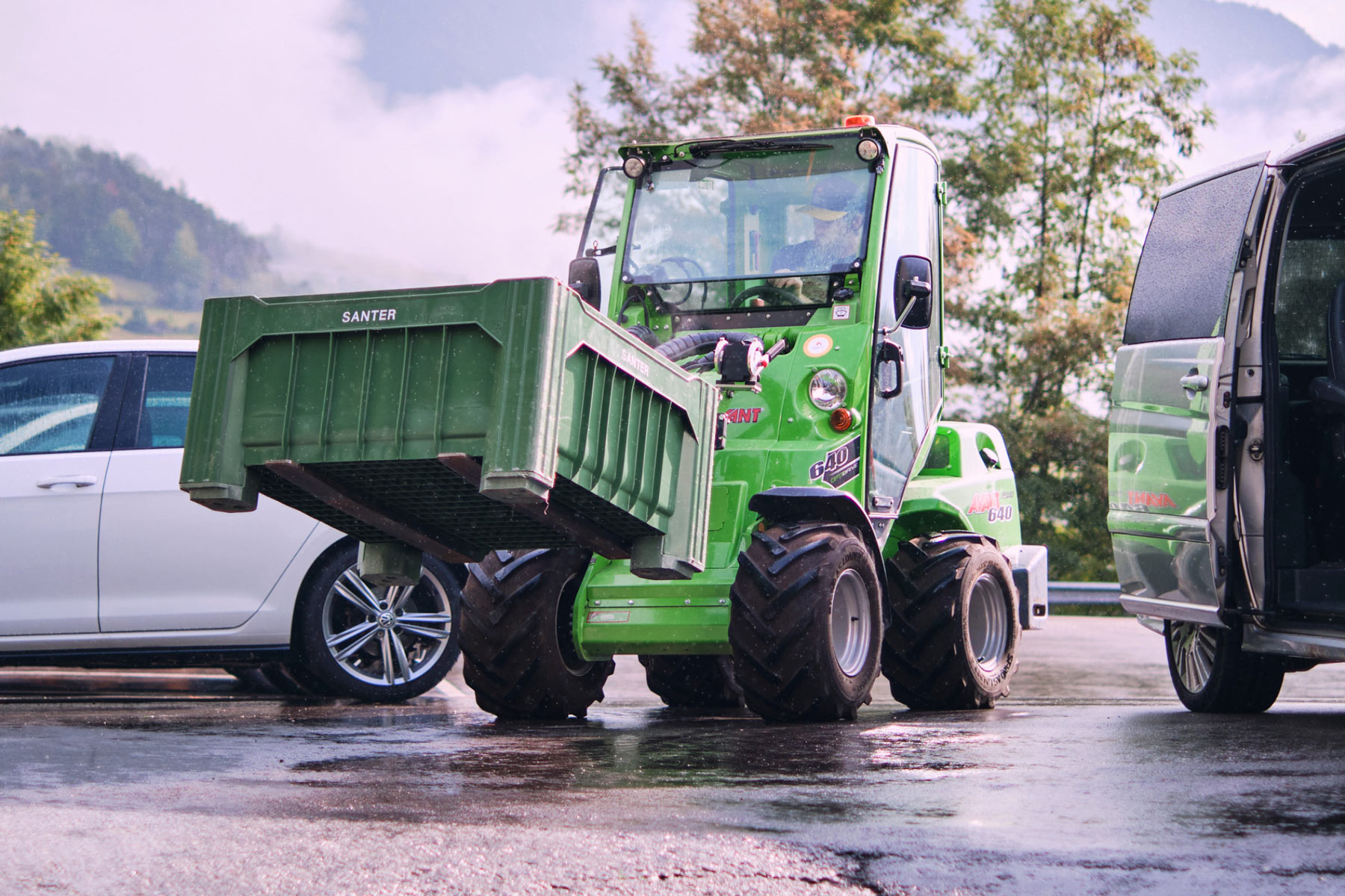 Grüner Gabelstapler mit Anhänger auf nassem Parkplatz, Berge im Hintergrund.