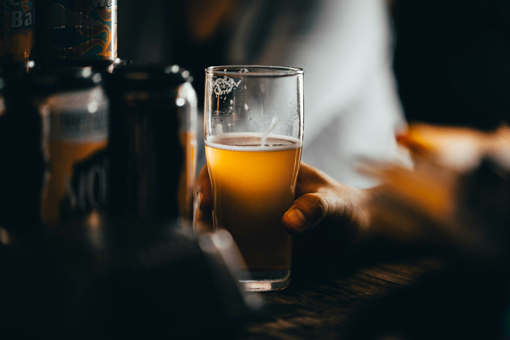 Hand hält ein Glas helles Bier in stimmungsvoller Bar-Atmosphäre